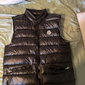 Moncler väst, öppen för byten - Säljer en snygg svart dunväst från Moncler. Västen har en design med dragkedja och två praktiska fickor med dragkedjor. Perfekt för kyliga höst- och vårdagar. Moncler-loggan är synlig på bröstet. Säljer då den var för liten men skit skön väst