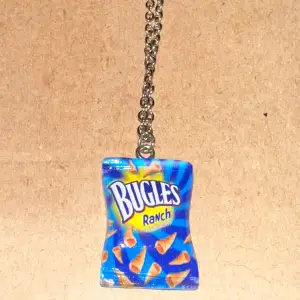 Ett unikt och roligt halsband med en miniatyr av en Bugles Ranch-chipspåse som hänge. Kedjan är i silverfärg och ger en cool kontrast till den färgglada påsen. Perfekt för dig som vill ha något annorlunda och lekfullt i din smyckessamling!