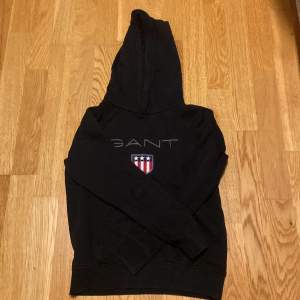 Säljer en svart hoodie från GANT med deras klassiska logga och ett coolt emblem på bröstet. Den har långa ärmar och en skön huva. Perfekt för en avslappnad stil! Det finns några små fläckar på ärmen, men annars i bra skick.