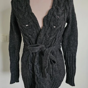 Mörkgrå stickad kofta från H&M - Säljer en supermysig mörkgrå stickad kofta från H&M. Den har ett snyggt flätmönster och ett bälte i midjan för en fin passform. Perfekt för höst och vinter! Koftan har långa ärmar och är i storlek XS. Perfekt att mysa i under kyliga dagar.