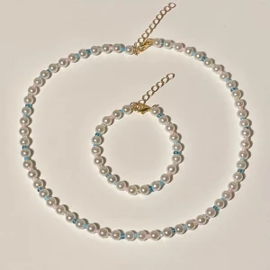 Pärlhalsband och armband med blå detaljer - Säljer ett superfint set med ett halsband och ett armband, båda med vita pärlor och små blå detaljer. Perfekt för att ge en elegant touch till vilken outfit som helst. Båda smyckena har justerbara kedjor i guld för att passa olika storlekar. Perfekt för fest eller sommarens alla tillfällen! ✨