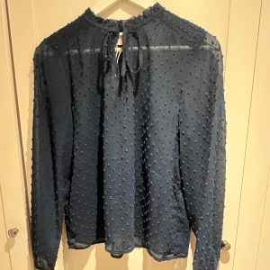 Mörkblå blus med knytband - Säljer en elegant mörkblå blus med knytband vid halsen. Blusen har ett diskret prickigt mönster och långa ärmar. Nyskick. Köparen står för frakten. Perfekt för både vardag och fest!
