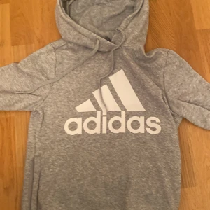 Grå hoodie från Adidas - Säljer en superbekväm grå hoodie från Adidas i storlek XS. Den har en klassisk design med Adidas-loggan tryckt i vitt på framsidan. Perfekt för både träning och mysiga dagar hemma. Huvan är justerbar med snören och materialet är mjukt och skönt.