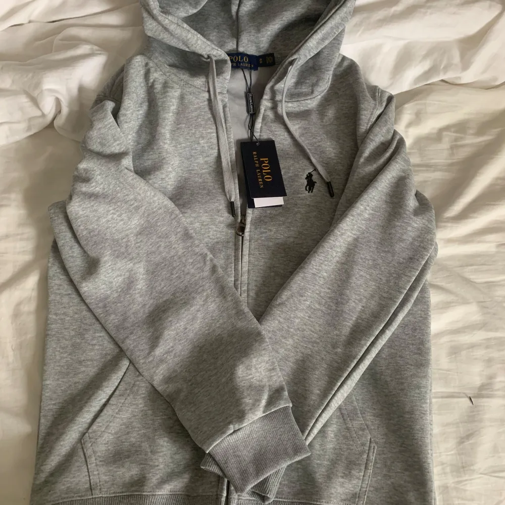 Säljer en snygg och bekväm grå hoodie från Polo Ralph Lauren. Den har en dragkedja framtill och den klassiska broderade loggan på bröstet. Perfekt för en avslappnad stil och passar till alla tillfällen. Huvan är justerbar med snören och det finns fickor på sidorna. Ett måste i garderoben!. Tröjor & Koftor.