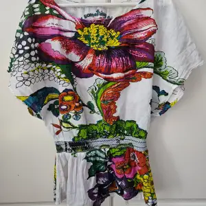 Säljer en superfin och färgglad topp från Desigual. Den har ett stort blommönster i rosa, rött, gult och grönt på en vit bakgrund. Toppen är kortärmad och har en lös passform, perfekt för sommardagar. Den är i bomull och känns väldigt bekväm att ha på sig.