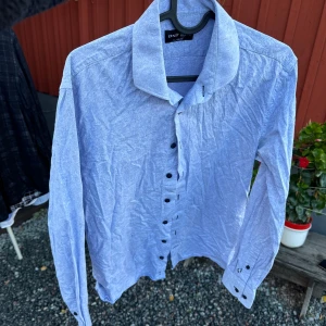 Blå skjorta från Ernst Alexis - Säljer en snygg blå skjorta från Ernst Alexis i storlek 39/40. Skjortan har ett diskret paisleymönster och är i slim fit. Den är långärmad med knappar längs framsidan och vid ärmsluten. Perfekt för både vardag och fest!