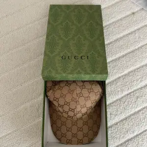 Säljer en snygg Gucci keps med det klassiska GG-monogrammet i brunt. Kepsen har en grön och röd rand på sidan. Insidan är fodrad med svart tyg och har Gucci-loggan tryckt runt om. Perfekt för att lyfta vilken outfit som helst!