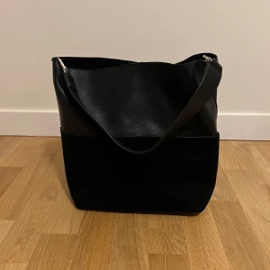 Svart Handväska  - Svart handväska från Åhléns, jättefint skick bara använd några gånger. Har två st. Yttre fack (där ”mockan” är). Säljer då jag fick den i present och den inte riktigt  passar min stil. Inköpspris: 599