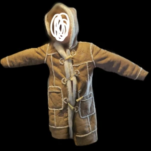 Brun pälsjacka med huva - Säljer en supermysig brun duffeljacka med huva. Den har långa ärmar och är dekorerad med vita sömmar och vackra knappar framtill. Perfekt för kalla vinterdagar! Storlek:128/8år