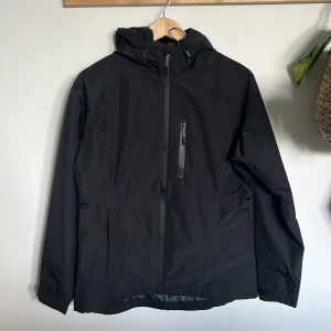 Svart vindjacka från McKinley - Säljer en svart vindjacka från McKinley i storlek 38. Jackan är perfekt för utomhusaktiviteter med sin vattentäta design (10,000 mm) och helt tejpade sömmar. Den är både vindtät och andas bra, vilket gör den idealisk för blåsiga och regniga dagar. Jackan har en dragkedja framtill och en praktisk huva.