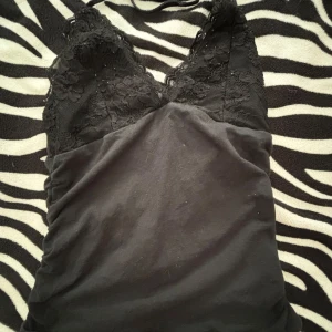 Svart linne med spetsdetaljer - Säljer ett snyggt svart linne med vackra spetsdetaljer. Toppen har en tight passform och är perfekt för festliga tillfällen. Den är ärmlös och har en fin spetsdesign upptill som ger en elegant touch.