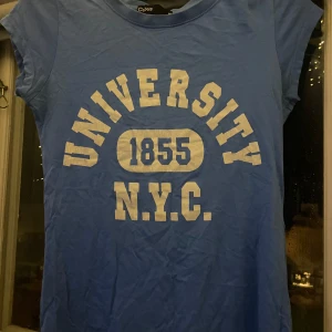 Blå t-shirt från Cubus med tryck - Säljer en blå t-shirt från Cubus med ett coolt tryck som säger 'University 1855 N.Y.C.'. Perfekt för en avslappnad stil och passar bra till jeans eller shorts. T-shirten är kortärmad och i bomull, vilket gör den skön att ha på sig.