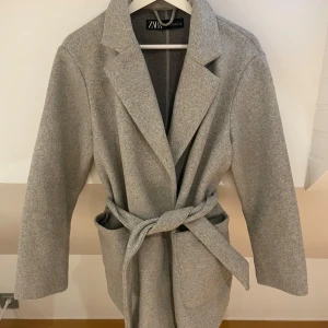 Grå kappa - Grå kappa från Zara med band runt midjan. Kan upplevas som textur/nopprig i tyget på kappan se bild 2, men skulle inte säga att de syns i helheten på kappan. Säljer då den inte används💓