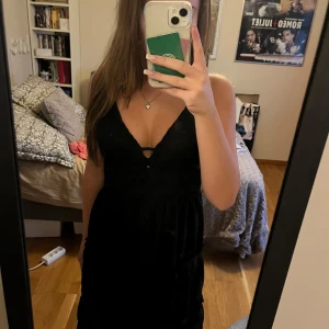 Svart klänning med spetsdetaljer - Säljer en elegant svart klänning med v-ringad design och spetsdetaljer på överdelen. Klänningen har en smickrande passform och är perfekt för festliga tillfällen. Den är ärmlös och har en fin struktur som ger en lyxig känsla.