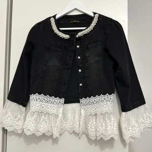 Svart jeansjacka med spetsdetaljer och pärlor - Säljer en unik svart jeansjacka från Neoma i storlek S/M. Jackan har långa ärmar och är dekorerad med vackra pärlor runt halsringningen. Den har också fina vita spetsdetaljer vid ärmslut och nederkant som ger en romantisk touch. Perfekt för att piffa upp vilken outfit som helst!