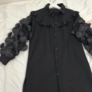 Svart klänning med unika ärmdetaljer från UMS Concept - Säljer en supercool svart klänning från UMS Concept. Den har en normal passform och är dekorerad med volanger längs bröstet och unika, cirkulära detaljer på ärmarna som verkligen sticker ut. Perfekt för en fest eller en kväll ute när du vill ha något annorlunda och stilrent. Klänningen är i nyskick och redo att bäras av någon som vill göra ett statement!