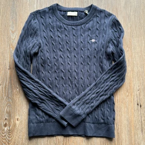 Mörkblå stickad tröja från GANT - Säljer en snygg mörkblå stickad tröja från GANT. Den har ett klassiskt flätmönster och en liten broderad logga på bröstet. Tröjan är långärmad och perfekt för höst och vinter. Supermysig och stilren!