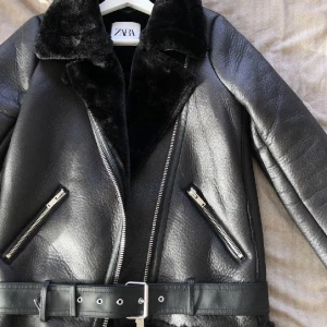Svart skinnjacka från Zara - Säljer en snygg svart skinnjacka från Zara med mjukt foder och pälskrage. Jackan har en cool bikerstil med dragkedjor och ett bälte i midjan. Perfekt för höst och vinter! Och jätte varm helt ny