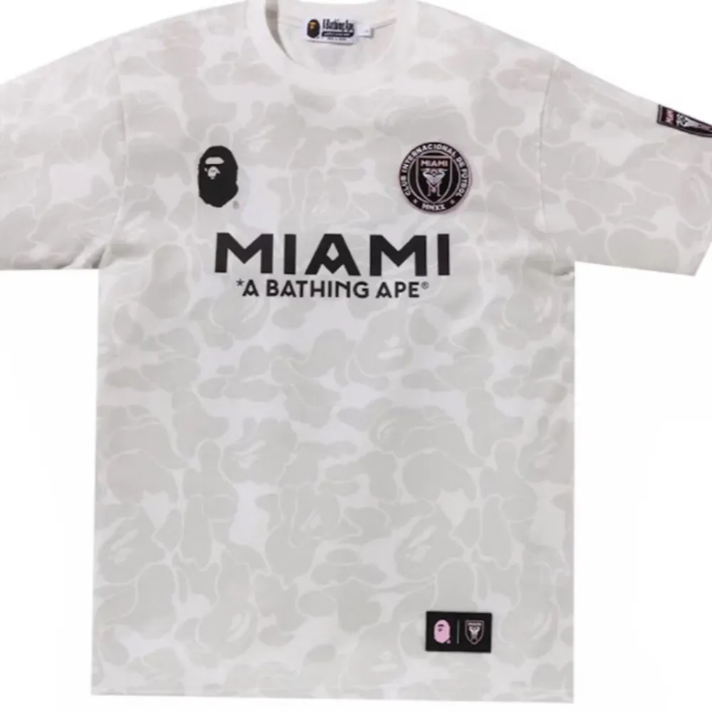 Säljer en supercool vit fotbollströja från samarbetet mellan A Bathing Ape och Miami. Tröjan har ett unikt kamouflagemönster och korta ärmar. Den har tryck med 'MIAMI' och 'A BATHING APE' på framsidan, samt loggor på bröstet och ärmen. . T-shirts.