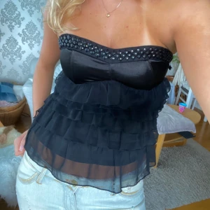 Svart axelbandslös topp med volanger - superfin svart axelbandslös topp med glittriga detaljer upptill och volanger! Perfekt för en utekväll eller fest! 