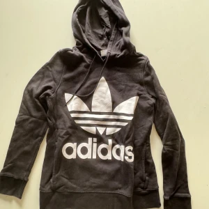 Svart hoodie från Adidas - Säljer en svart hoodie från Adidas med den klassiska loggan i vitt på framsidan. Den har en bekväm huva och långa ärmar. Perfekt för en avslappnad stil eller träning. Passar bra till höst och vinter!