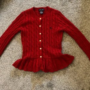 Röd stickad kofta från Ralph Lauren - Säljer en superfin röd stickad kofta från Ralph Lauren. Den har ett klassiskt flätmönster och knappar framtill. Koftan är långärmad och har en söt volangdetalj vid nederkanten. Perfekt för höst och vinter!