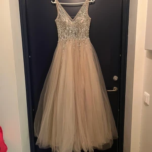 Champagnefärgad balklänning med med paljetter och stenar - Elegant champagnefärgad långklänning med glittrande paljetter på överdelen och en v-ringad design. Klänningen har en öppen rygg och ett flödande tyllkjol som ger en drömlik känsla. Perfekt för speciella tillfällen, som typ balen! Klänningen kostar ca 5500 i nypris.