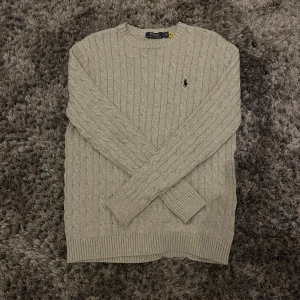 Grå stickad tröja från Ralph Lauren - Säljer en stilren grå stickad tröja från Ralph Lauren med kabelstickat mönster. Tröjan har långa ärmar och en broderad logotyp på bröstet. Perfekt för kyliga dagar!