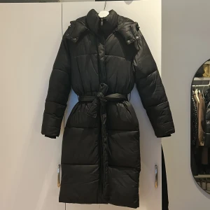Svart dunjacka med bälte - Säljer en jättefin svart lång vinterjacka med bälte i midjan. Jackan har en hög krage och avtagbar luva. Dragkedja framtill och praktiska fickor på sidorna.