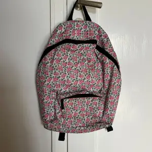 Snygg ryggsäck med ett blommigt mönster i rosa och svart på vit bakgrund. Väskan har flera praktiska fack med dragkedjor och justerbara axelremmar. Perfekt för att bära med sig det nödvändigaste med stil.