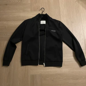 Svart zip tröja från Les Deux - Snygg svart tröja från Les Deux med dragkedja framtill och broderad logga på bröstet. Tröjan har långa ärmar och ribbade muddar vid ärmslut och nederkant. Perfekt för en stilren look. Köpt för 1100kr och är som ny, sparsamt använd. Mycket bra skick!