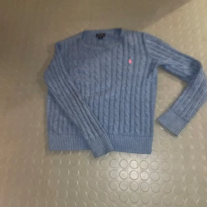 Blå stickad tröja från Polo Ralph Lauren - Säljer en snygg blå stickad tröja från Polo Ralph Lauren med kabelstickat mönster. Tröjan har långa ärmar och en liten rosa logga på bröstet. Perfekt för kyliga dagar! Passar dig som är ca 172-160