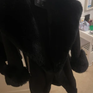 Svart pälsjacka med bälte - Elegant svart jacka med mjuk pälsdetalj på krage och ärmslut. Jackan har en stilren design med dragkedja och ett bälte i midjan för en snygg passform. Den är i storlek XS/S (pris kan diskuteras eller ange prisförslag)