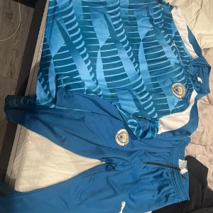 Blå fotbollströja och byxor från Puma - Snygg blå fotbollströja med matchande byxor från Puma. Tröjan har ett coolt mönster i olika blå nyanser och vita detaljer. Perfekt för fotbollsfans som vill ha en stilren look på och utanför planen.