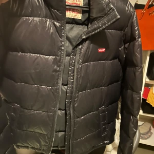 Svart dunjacka från Levi's st s - Snygg svart dunjacka från Levi's med dragkedja framtill. Jackan har en klassisk pufferdesign och är perfekt för kyligare väder. Levi's logga syns på bröstet..Jackan är perfekt för lite mildare dagar