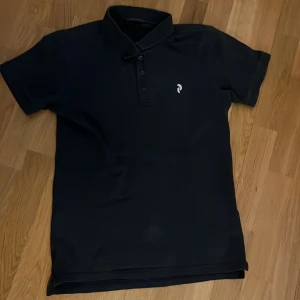 Svart pikétröja med broderad logga - Snygg svart pikétröja med en diskret broderad logga på bröstet. Tröjan har en klassisk krage och knappar framtill. Perfekt för en stilren look.