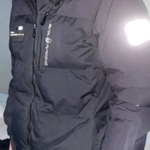 Svart dunjacka från Sail Racing - Snygg svart dunjacka från Sail Racing med dragkedja och logotyp på bröstet. Jackan har en pufferdesign och är perfekt för kyligare väder. Den har långa ärmar och en praktisk huva.