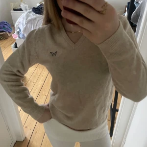 Beige tröja med broderad detalj - Snygg beige stickad tröja med v-ringning och en liten broderad detalj på bröstet. Tröjan har långa ärmar och ribbade muddar vid ärmslut och nederkant. Perfekt för en stilren look.