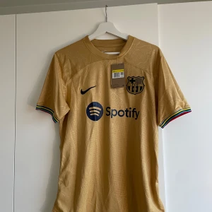 Barcelona fotbollströja från Nike - Snygg guld fotbollströja från Nike med korta ärmar och klubbmärke på bröstet. Tröjan har färgglada detaljer vid ärmsluten och sponsorer som Spotify och UNHCR tryckta på framsidan och baksidan. FC Barcelona bortatröja 22/23 Perfekt för fotbollsfans! 