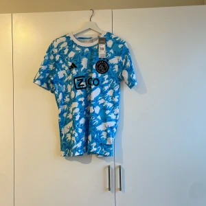 Blå och vit fotbollströja från Adidas - Snygg fotbollströja från Adidas i blått och vitt med ett unikt mönster. Tröjan har korta ärmar och en rund halsringning. Perfekt för fotbollsfans som vill sticka ut på planen eller läktaren. Ajax special edition tröja 23/24