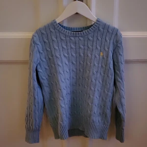 Ljusblå stickad tröja från Ralph Lauren - Säljer en ljusblå stickad tröja från Ralph Lauren med kabelstickat mönster. Tröjan har långa ärmar och en liten gul broderad logotyp på bröstet. Perfekt för kyliga dagar!
