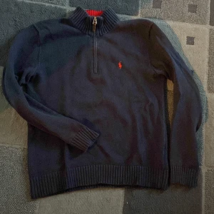 Mörkblå tröja från Polo Ralph Lauren - Snygg mörkblå tröja från Polo Ralph Lauren med röd broderad logga på bröstet. Tröjan har en dragkedja vid halsen och en ribbad krage i rött. Tröjan är i storlek s och har lite tecken på användning!