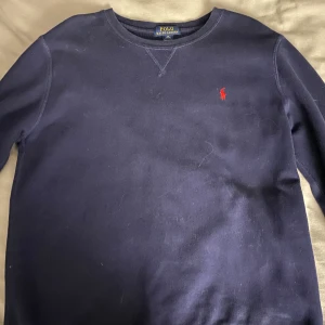 Mörkblå tröja från Ralph Lauren - Säljer en stilren mörkblå tröja från Ralph Lauren med det klassiska röda logotypbroderiet på bröstet. Tröjan har rund hals och långa ärmar, perfekt för en avslappnad look.