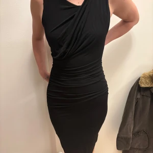 Svart klänning från Tiger of Sweden - Elegant svart klänning från Tiger of Sweden med draperad design och smickrande passform. Klänningen har en dragkedja baktill och är ärmlös, vilket ger en stilren look. Perfekt för en kväll ute eller en speciell tillställning.