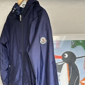 Blå vindjacka från Moncler - Vindjacka från Moncler, modell Lepontine Reversible, i storlek M. Jackan är endast testad och i nästintill nyskick. Vändbar design med en sida i stilren mörkblå färg och en sida med ett snyggt Moncler-mönster, vilket gör den både mångsidig och trendig. Perfekt för vår- och höstbruk! Kvaliteten är som förväntat från Moncler – toppklass. Kvitto finns. Hör gärna av dig vid frågor!