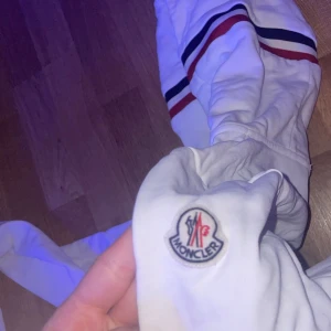 Vit hoodie från Moncler - Säljer en snygg vit hoodie från Moncler med dragkedja och randiga detaljer i rött och blått på huvan. Perfekt för en stilren och avslappnad look. Moncler-logga på bröstet ger en exklusiv touch. Har snöre till konkatka mig för mer bilder