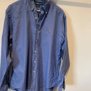 Blå skjorta från Ralph Lauren - Snygg blå skjorta från Ralph Lauren i 100% bomull. Skjortan har långa ärmar och klassiska knappar framtill. Den är dekorerad med ett broderat märke  på bröstet. Perfekt för en stilren look. (Pris går att diskutera)