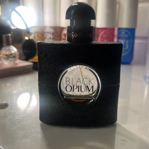 Black Opium parfym från Yves Saint Laurent - Säljer en elegant och mystisk parfymflaska av Black Opium från Yves Saint Laurent. Flaskan är svart med en glittrande yta och har en rund genomskinlig del i mitten där namnet syns. Perfekt för den som älskar en djärv och sensuell doft.