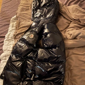Svart glansig dunjacka från moncler (barn) - Säljer en svart Maya dunjacka från moncler med luva och dragkedja. Jackan har en quiltad design och är perfekt för kyligare väder, den är dock skadad på en av fickorna och på insidan men man tänker ej på det