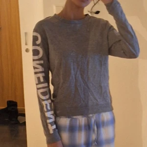 Grå tröja med text på ärmen - Säljer en grå tröja med texten 'CONFIDENT' tryckt längs ena ärmen. Tröjan har en rund hals och långa ärmar, perfekt för en avslappnad stil. Är i storlek XS